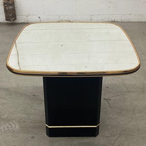 May include: Square side table with a mirrored top and a black base. The table has a gold trim around the mirrored surface and at the base of the pedestal. The table is approximately 24 inches wide and 24 inches long.