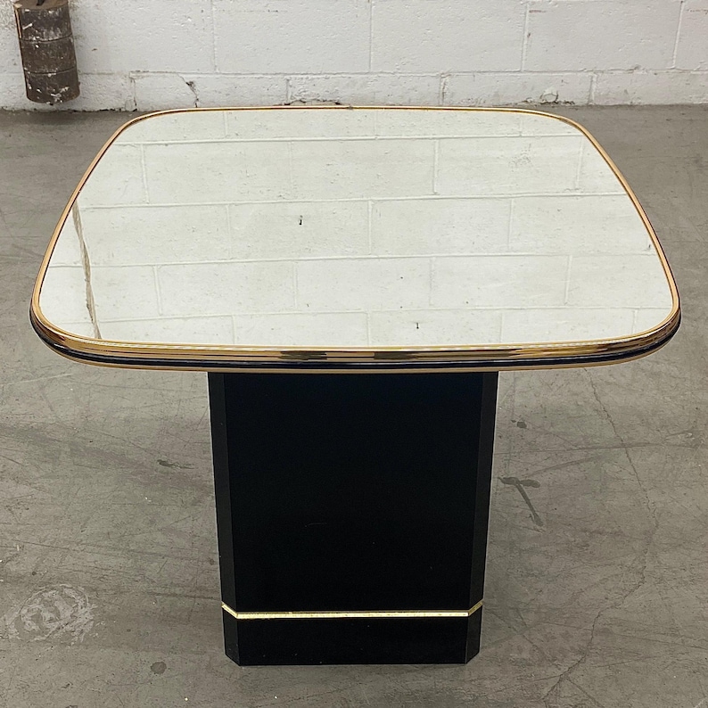 May include: Square side table with a mirrored top and a black base. The table has a gold trim around the mirrored surface and at the base of the pedestal. The table is approximately 24 inches wide and 24 inches long.