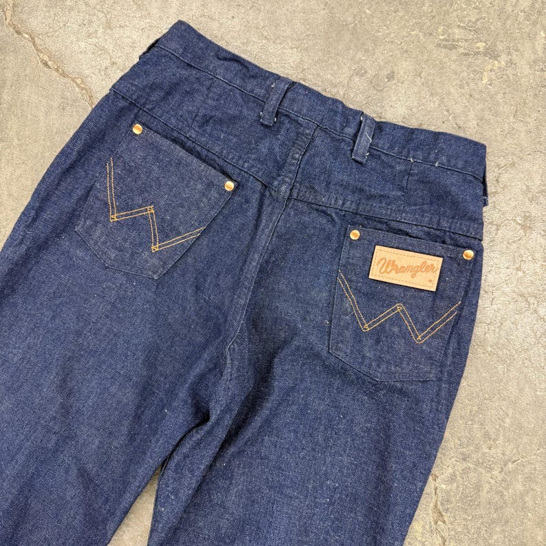 May include: Dark blue denim Wrangler jeans. The jeans have gold stitching on the back pockets and a Wrangler label. The jeans have gold rivets and a classic five-pocket design.