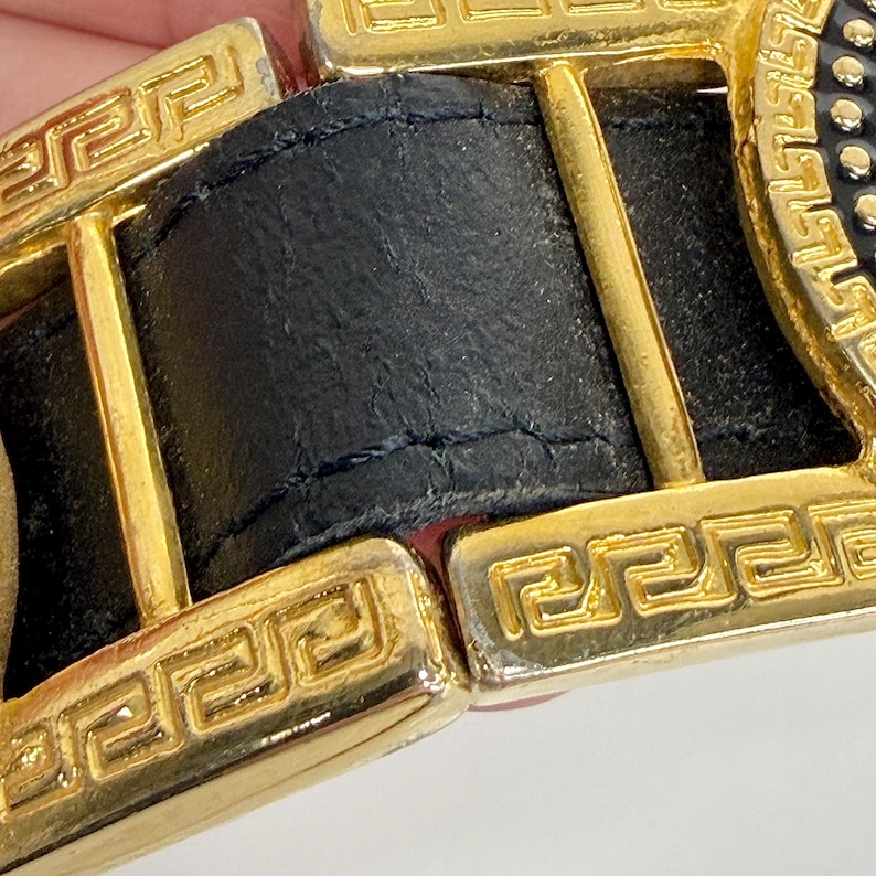 Puede incluir: Un cintur&oacute;n de cuero negro con una prominente hebilla de metal dorado. La hebilla es rectangular con bordes redondeados y muestra un patr&oacute;n de greca grabado a lo largo de su borde. La correa de cuero negro pasa a trav&eacute;s del marco met&aacute;lico.