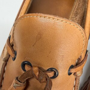 May include: A tan leather boat shoe featuring a moccasin-style toe construction with visible contrast stitching. The shoe includes dark metal eyelets and a leather lace threaded through the sides and tied at the front vamp area.