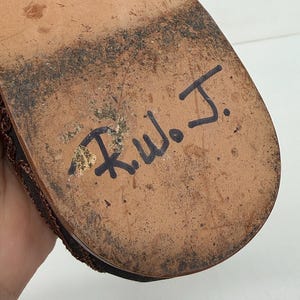 May include: Close-up of a brown shoe sole with the handwritten initials "R.W.J." in black ink. The sole is worn, showing signs of use, with a textured surface and dark patches.