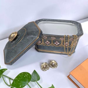 May include: A rectangular dark gray lidded box with a gold-toned knob and gold trim. The exterior features a repeating floral pattern in pink and gold. A thin gold chain hangs over the edge, and two gold-toned flower-shaped earrings sit nearby.