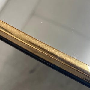 May include: Close-up of a gold-colored metal frame with a dark blue edge. The frame has a brushed texture and appears to be part of a mirror or picture frame. The gold color has a slight sheen, reflecting the light.