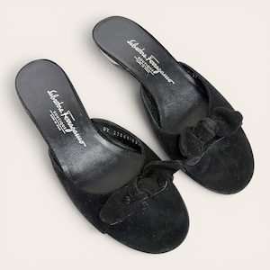 May include: A pair of black suede mules featuring a rounded toe and a decorative bow detail on the front. The interior footbed is black and displays the white text Salvatore Ferragamo Boutique Made in Italy with a serial number visible.