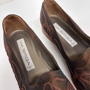 May include: Close-up of two Via Spiga flats. The shoes are dark brown with intricate embroidered detailing in a lighter brown. The interior is a lighter brown with a label that reads "VIA SPIGA MADE IN ITALY".