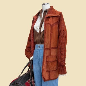 May include: A rust-colored suede and knit jacket with a brown button-up shirt and blue jeans. The jacket has a patchwork design and cable knit sleeves. A black and red duffel bag is held in the left hand.