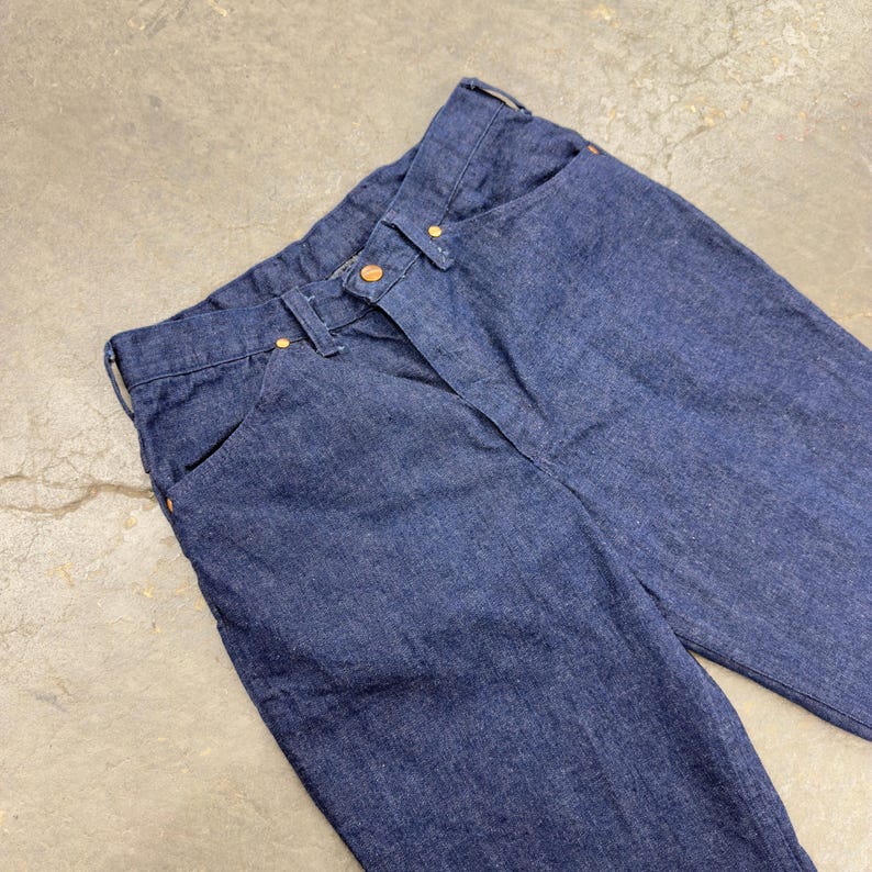 May include: Close-up of dark blue denim jeans. The jeans feature a classic five-pocket design with copper-coloured rivets and a button closure. The fabric appears to be a medium-weight denim.