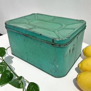 May include: A rectangular mint green metal storage tin with a geometric embossed lid. The container features visible signs of wear, including surface scratches and minor rust along the edges, and is positioned near three yellow lemons.