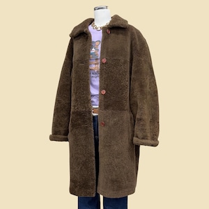 May include: A long, brown shearling coat with a wide collar and three large buttons. The coat is worn over a purple t-shirt with text and blue jeans. The coat has a relaxed fit and a soft, textured appearance.