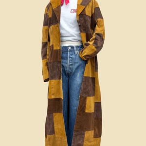 May include: A long patchwork coat made of alternating brown and mustard yellow corduroy panels. Underneath, a white t-shirt with red text, blue denim jeans, and a bright red neckerchief are displayed on a mannequin torso.