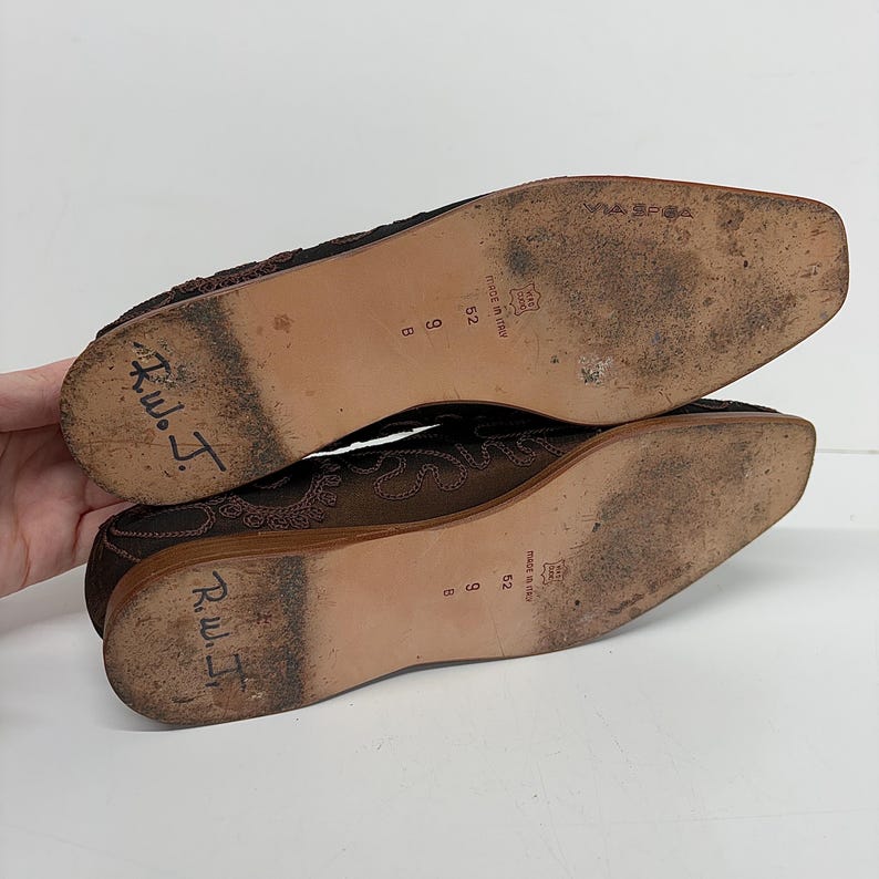 May include: A pair of brown Via Spiga flats, size 9 B, with decorative stitching. The soles show wear and tear, with the inscription "R.W.J." written on the sole of each shoe. Made in Italy.