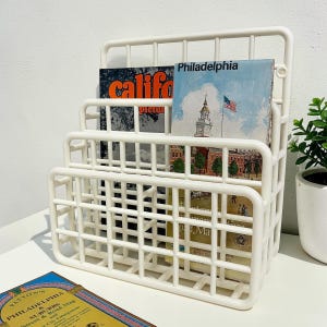 May include: A white, three-tiered magazine rack filled with books and magazines. The rack has a grid-like design and is made of plastic. The books include titles like "California Pictures" and "Philadelphia". A small potted plant is visible on the right.