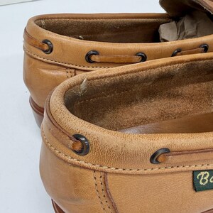 May include: A pair of tan leather loafers featuring a moccasin-style construction with dark metal eyelets and a thin leather lacing detail around the collar. The interior is lined with soft suede, and a small green fabric tag is visible on the side.