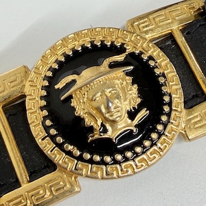 Puede incluir: Un cintur&oacute;n de cuero negro con una gran hebilla circular dorada. La hebilla tiene una incrustaci&oacute;n central de esmalte negro con un relieve dorado de una figura alada, rodeada por un borde con patr&oacute;n de greca y eslabones de metal dorado.