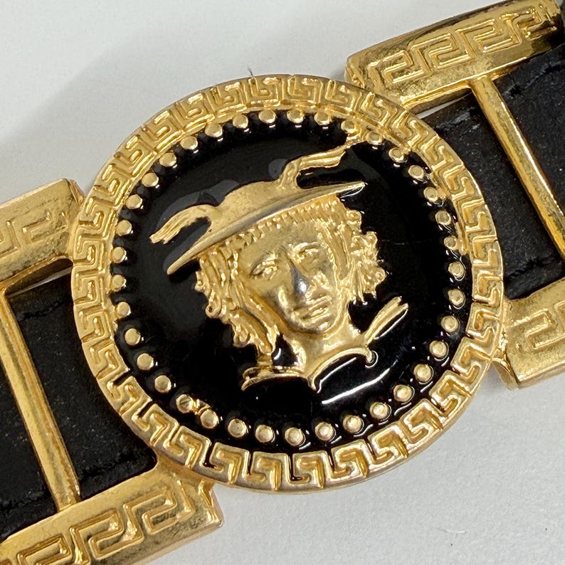 Puede incluir: Un cintur&oacute;n de cuero negro con una gran hebilla circular dorada. La hebilla tiene una incrustaci&oacute;n central de esmalte negro con un relieve dorado de una figura alada, rodeada por un borde con patr&oacute;n de greca y eslabones de metal dorado.
