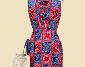May include: A sleeveless dress featuring a red and blue bandana-style patchwork print with a ruffled collar detail. The dress has a fitted bodice and a flared skirt. A plain off-white canvas tote bag is positioned next to the dress on the left side.