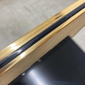 May include: Close-up of a gold-colored picture frame with a black border. The frame has a shiny, reflective surface and a black backing. The frame is angled, showing the side profile.
