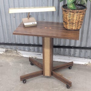 May include: A rolling side table with a dark wood grain finish, a central column featuring light-colored cane webbing, and a four-pronged base with casters. A gold-toned desk lamp and a woven wicker basket containing a green plant sit on top.