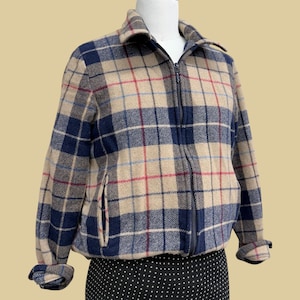 May include: A full-zip jacket in a plaid pattern. The jacket features a beige base with navy blue, red, and light blue plaid lines. It has a collared neckline, a front zipper, and two side pockets. The cuffs are rolled up.