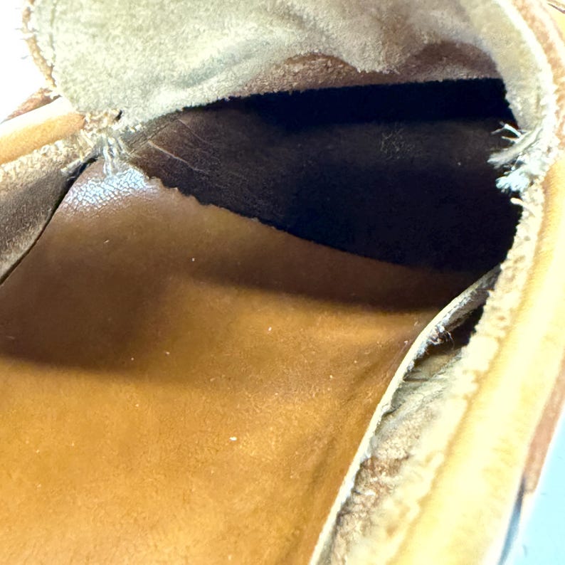 May include: A tan leather loafer shoe showing the interior construction. The shoe features a smooth brown leather insole, a dark brown leather lining along the heel area, and visible stitching along the top edge of the opening.