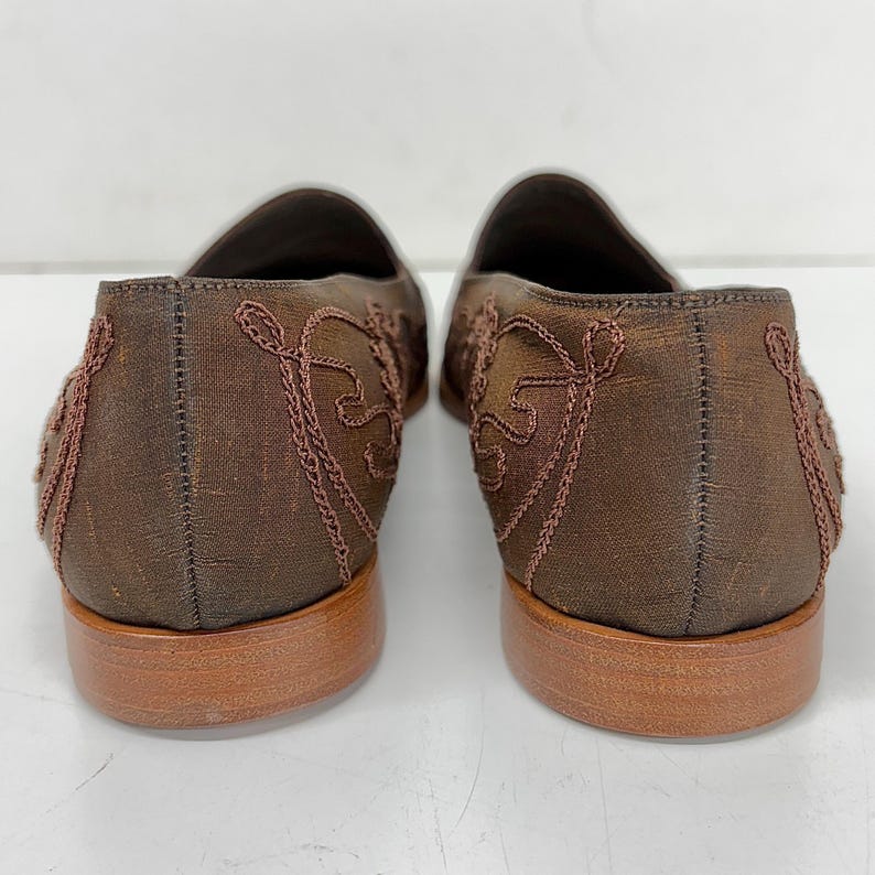 May include: A pair of brown loafers with embroidered detailing. The shoes have a low heel and a decorative pattern on the sides. The color of the shoes is a dark brown with a lighter brown sole.