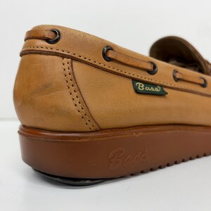 May include: A tan leather boat shoe featuring a dark brown rubber platform sole with the Bass logo embossed on the side. The shoe includes a leather lace detail threaded through dark metal eyelets and a small green tag with the Bass brand name.