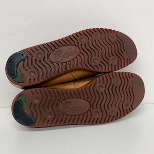 May include: A pair of tan leather shoes with brown rubber outsoles featuring a wavy tread pattern, circular indentations, and a black heel insert. The word Bass is embossed in the center of each sole, showing the bottom view of the footwear.