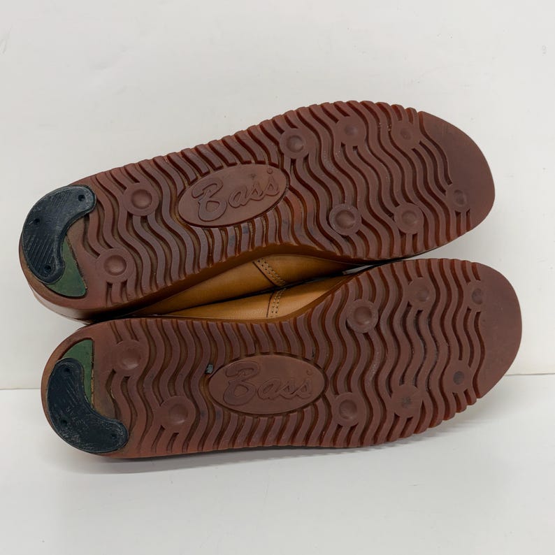 May include: A pair of tan leather shoes with brown rubber outsoles featuring a wavy tread pattern, circular indentations, and a black heel insert. The word Bass is embossed in the center of each sole, showing the bottom view of the footwear.