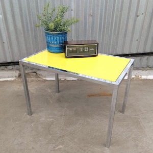 May include: A rectangular side table with a bright yellow top and a silver-toned metal frame. A blue textured ceramic pot containing a green plant and a dark brown rectangular flip clock displaying the numbers 1 and 22 sit on the table surface.