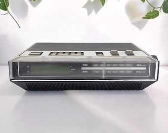 May include: A black General Electric AM/FM clock radio with a digital display, analog tuning dial, and top-mounted control buttons. The front panel features frequency scales for FM and AM bands, with clear text labels for station tuning and clock settings.