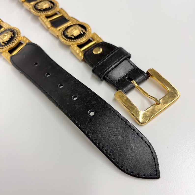 Puede incluir: Un cintur&oacute;n de cuero negro con una serie de medallones circulares de metal dorado con centros negros, cada uno con una figura dorada en relieve. El cintur&oacute;n incluye una hebilla de metal dorado y un extremo puntiagudo con cinco agujeros.