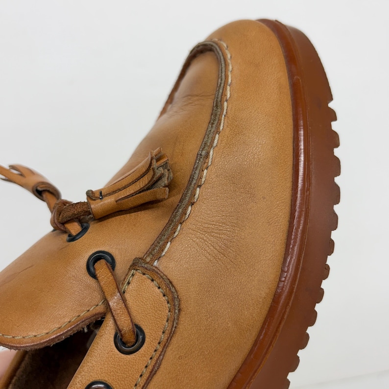 May include: A tan leather loafer featuring a moccasin-style stitched toe, a decorative leather tassel, and black metal eyelets for the lacing. The shoe is constructed with a thick, dark brown rubber lug sole with a prominent tread pattern.