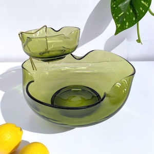May include: A two-tiered olive green glass serving dish featuring a large, wide bowl at the base and a smaller, matching bowl suspended above it by a thin gold-toned metal wire frame. The glass has a smooth, wavy, and organic rim design.