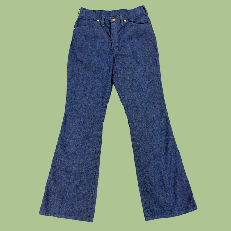 May include: Blue denim bell-bottom jeans. The jeans have a high waist, a button and zip closure, and classic five-pocket styling. The denim is a medium wash, and the legs flare out from the knee.