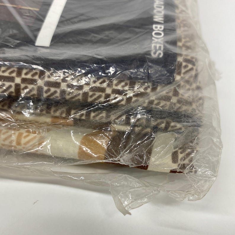 May include: A close-up of folded fabric in a clear plastic bag. The fabric has a repeating geometric pattern in shades of brown, beige, and white. The text "SHADOW BOXES" is partially visible on a dark background.