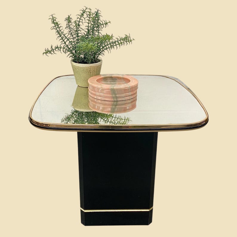May include: A black side table with a mirrored top and gold trim. A small potted plant and a decorative pink and gray stone ashtray sit on the table. The table has a rectangular base.