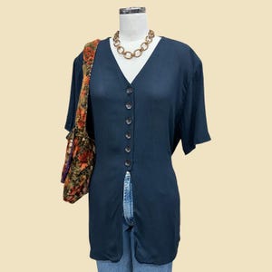 May include: A dark navy blue button-down shirt with a V-neck and short sleeves. The shirt is paired with blue jeans and a gold chain necklace. A shoulder bag with a floral pattern is on the left.