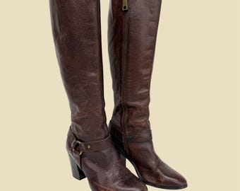 May include: A pair of brown leather knee-high boots. The boots have a pointed toe, a side zipper, and a buckle detail around the ankle. The heels are approximately 3 inches high.