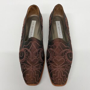May include: A pair of Via Spiga brown satin loafers with intricate embroidered detailing. The shoes have a square toe and a low heel. The interior of the shoe is a light tan colour.