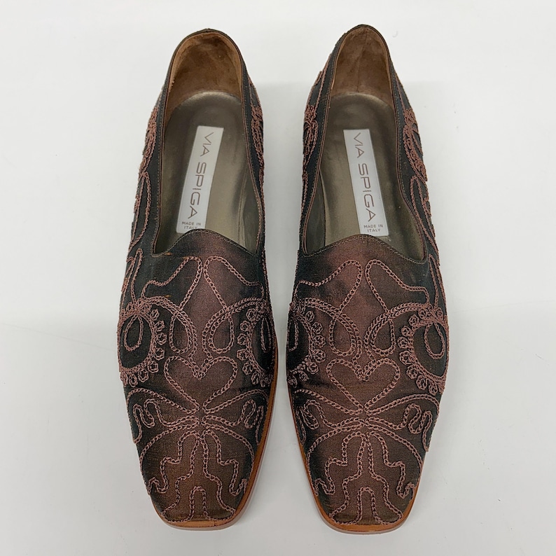 May include: A pair of Via Spiga brown satin loafers with intricate embroidered detailing. The shoes have a square toe and a low heel. The interior of the shoe is a light tan colour.