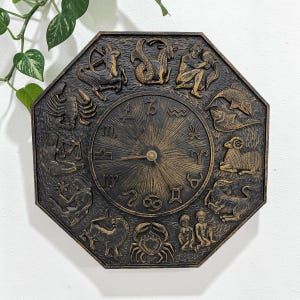 May include: An octagonal wall clock with a bronze finish, featuring embossed zodiac symbols around the perimeter. The clock face has a sunburst design with gold-colored hands. The clock is mounted on a white wall.