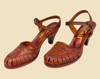 May include: A pair of vintage brown leather sandals with a heel. The sandals have an ankle strap with a buckle, a decorative stitched design on the toe, and the word "SENAS" printed in gold. The heels are dark brown.