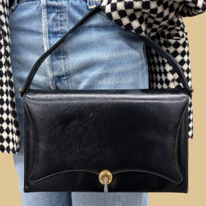 May include: A black structured handbag with a textured finish, featuring a single top handle and a gold-toned circular clasp at the center of the front flap. The bag has a curved, scalloped edge design along the bottom of the flap closure.