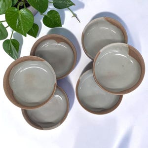 May include: A set of five small, round ceramic bowls with a natural, unglazed terracotta exterior and a smooth, glossy light gray interior glaze. The bowls are arranged in a cluster, showcasing their circular shape and contrasting textures.