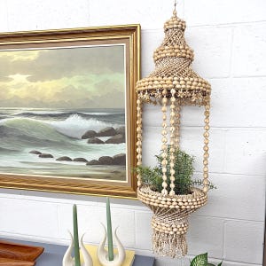 May include: A hanging seashell planter with a plant inside. The planter is made of various sizes of seashells and has a decorative, tiered design. The planter is hanging in front of a white brick wall. A framed painting of a seascape is in the background.
