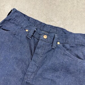 May include: Close-up of dark blue denim jeans. The jeans feature a button closure and brass-coloured rivets. The fabric shows signs of wear, with slight fading and creasing. The jeans are folded, showing the waistband and front pockets.