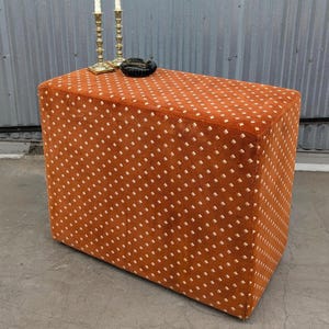 May include: A rectangular ottoman upholstered in burnt orange fabric with a repeating white diamond pattern. Two brass-colored metal candlesticks and a small black circular dish are positioned on the flat top surface of the furniture piece.