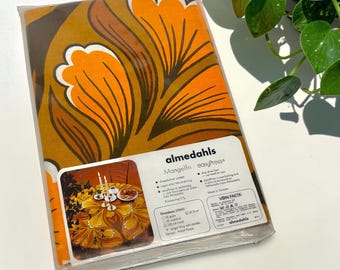 May include: A packaged round tablecloth featuring a bold, large-scale floral pattern in shades of orange, brown, and white. The packaging includes the Almedahls brand name, product details, and a small inset photograph showing the tablecloth in use.