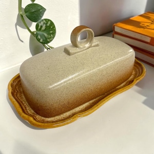 May include: A rectangular ceramic butter dish with a speckled cream-colored lid featuring a central circular handle. The base has a scalloped edge and a gradient finish that transitions from a light cream to a deep, warm brown at the bottom rim.